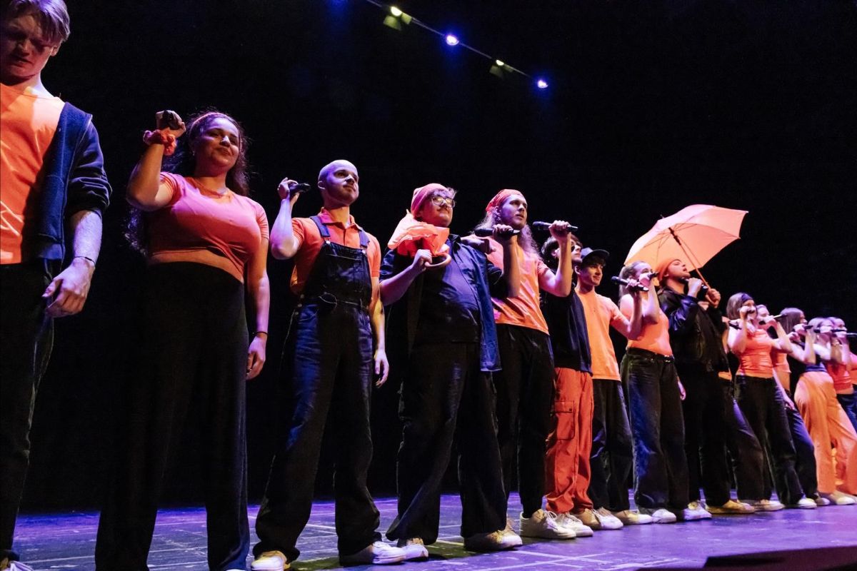 A group of people stand in a line on a stage. they are wearing orange clothes and they are holding hands. 