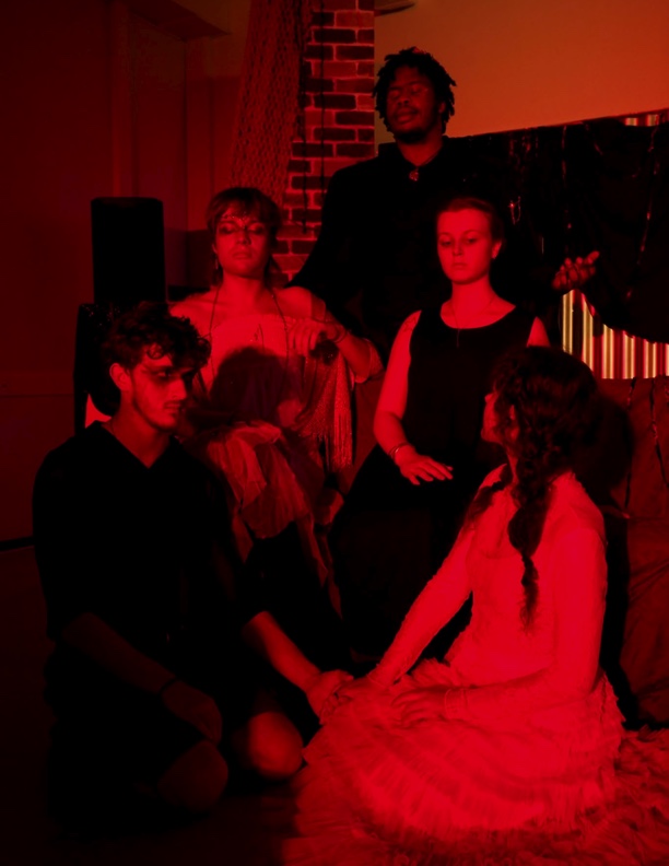 Five people dressed in blacks and whites sit on a stage looking at each other. They are bathed in red light. 