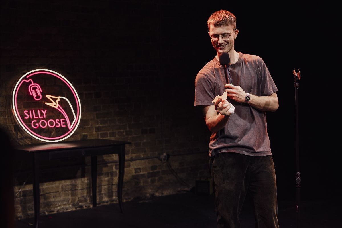 A man wearing a purple shirt stands on a stage with a mic. There is a light up sign behind them that says Silly Goose!