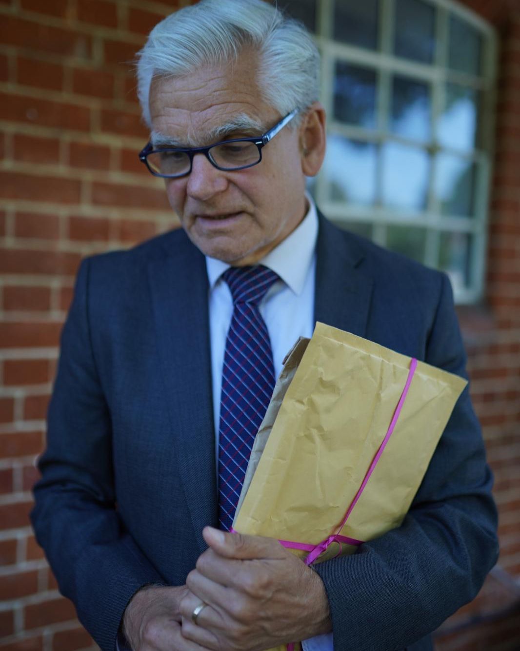 An old man with white hair holds a package and looks down.
